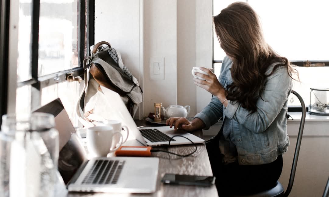 Woman with a cup of coffee working on a laptop beside a sunlit window